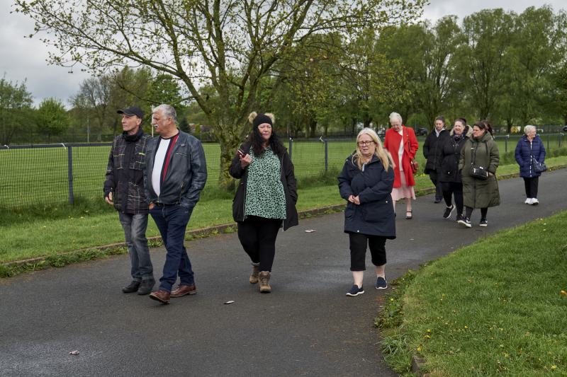 Mae grwp o bobl yn cerdded ar hyd llwybr y parc, o amgylch ysgafn a choed. Maent wedi gwisgo'n gynnes, yn awgrymu diwrnod oer, gan drosglwyddo awyrgylch ysgafn a chymdeithasol.