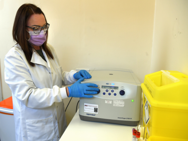 Principal investigator with long brown hair using blood spinning machine wearing a white coat, blue gloves and a mask