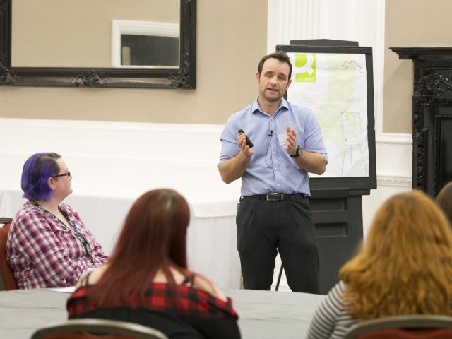 Male wearing a blue shirt and brown hair speaking at a conference in front of an audience