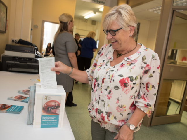 An elderly woman in flowery short putting a piece of paper in a consent box.