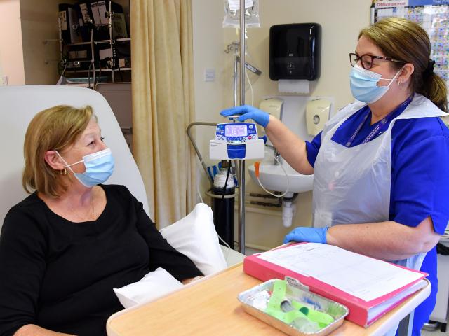 A female nurse in blue scrubs assisting a female patient in a black long-sleeved shirt.