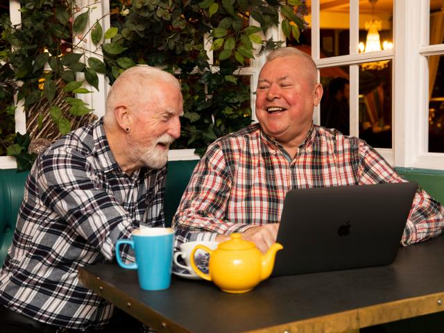 Two old men laughing at memes on a macbook