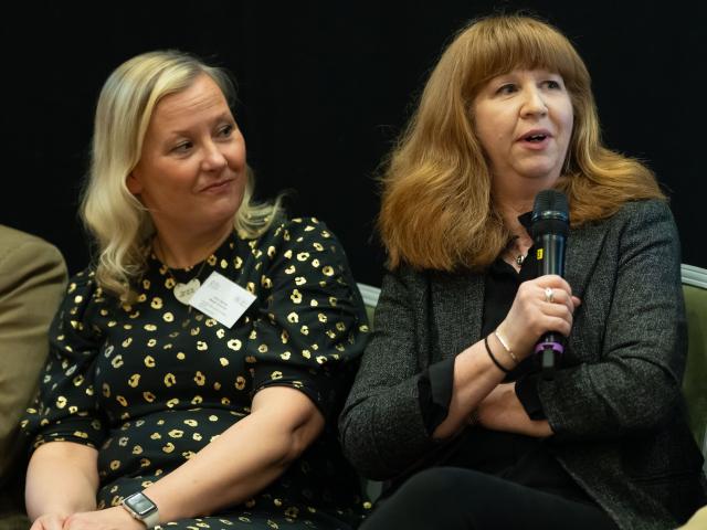 Two women sat on chairs on a stage with one speaking into a microphone