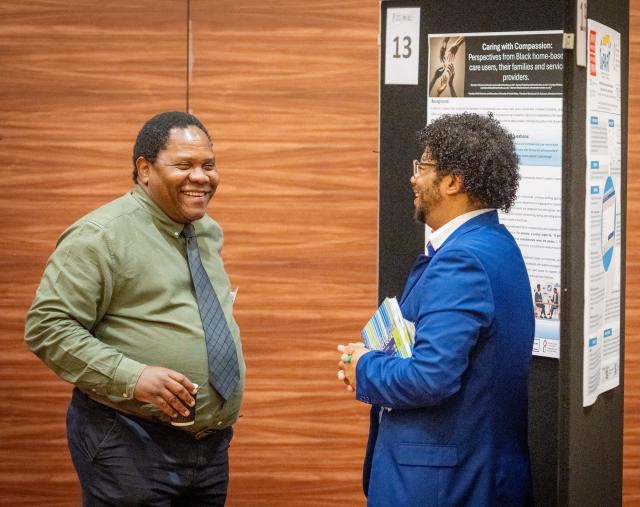 Two men stand and converse in front of a research poster at a conference; one holds a drink and the other carries folders.