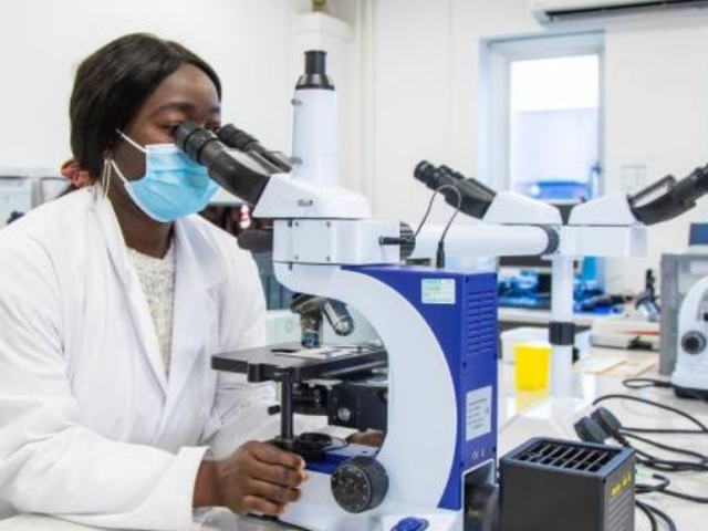 a_woman_using_a_microscope_wearing_lab_coat_and_face_mask