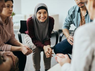 A group of people sitting in a circle with a focus on a young woman wearing a hijab. 