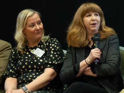Two women sat on chairs on a stage with one speaking into a microphone