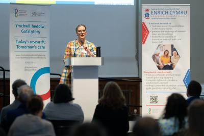 A person stands at a podium giving a presentation to an audience, with informational banners and a screen displayed behind them.